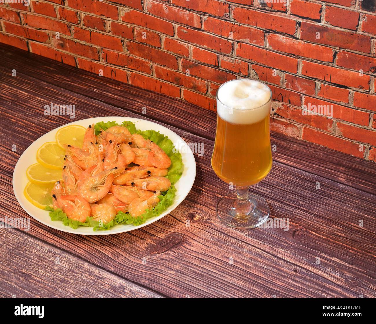 A tall glass of light beer with foam and a plate with boiled shrimp and lemon slices on a wooden table. Close-up. Stock Photo