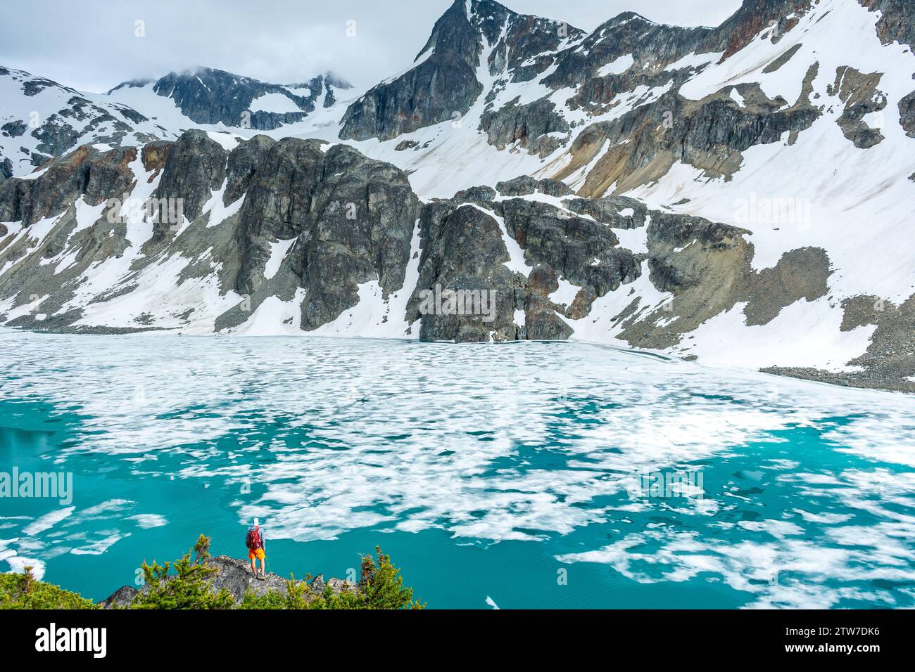 Lone hiker gazes across the icy expanse of Wedgemount Lake beneath towering cliffs. Stock Photo
