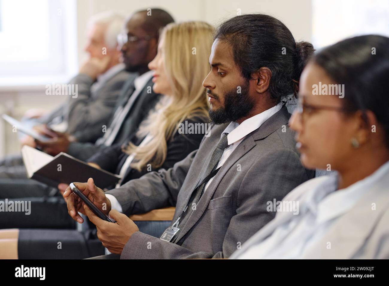 Young businessman in suit using mobile phone while sitting at conference with other people Stock Photo