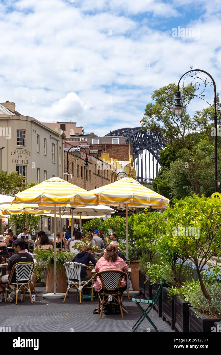 Outdoor dining under yellow and white striped umbrellas in The Rocks, with historic architecture and Sydney Harbour Bridge in the background. Stock Photo