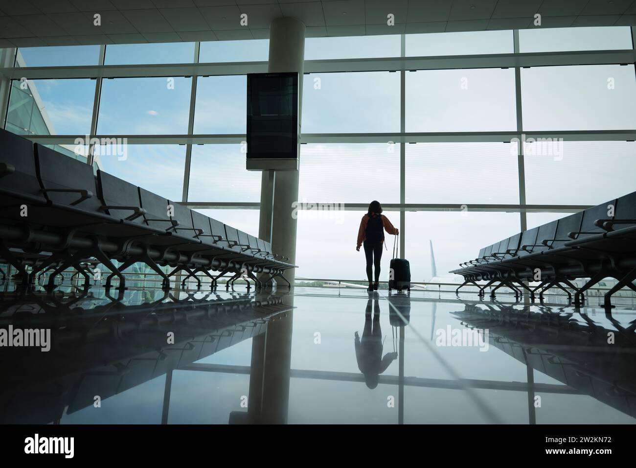Woman with suitcase in hand, looks out the airport window while waiting for her flight. Stock Photo