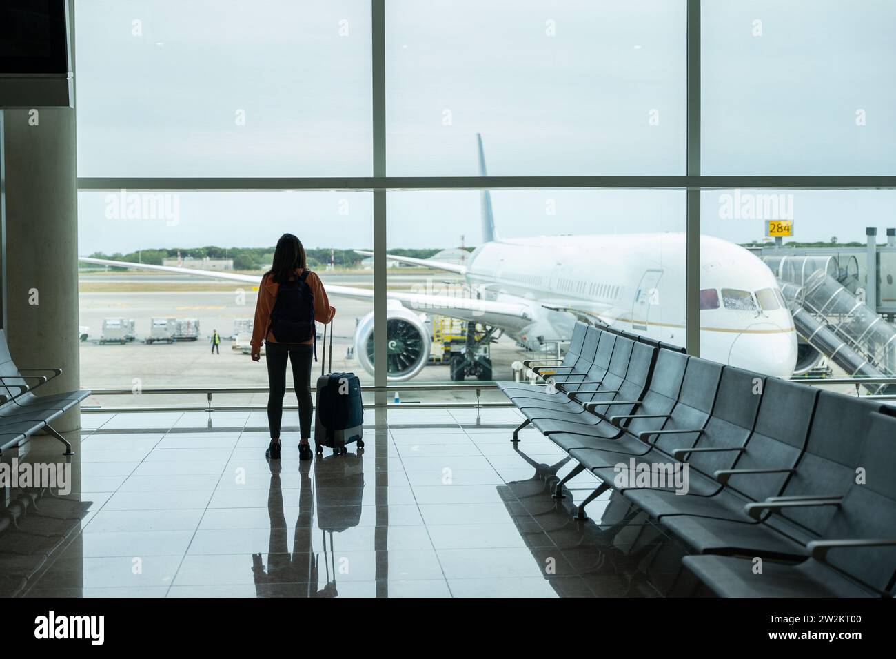Woman with suitcase in hand, looks out the airport window while waiting for her flight. Stock Photo