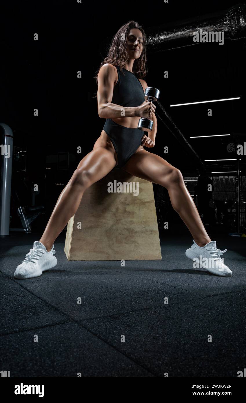 Tall athletic woman posing in the gym on a bench with dumbbells. Biceps ...