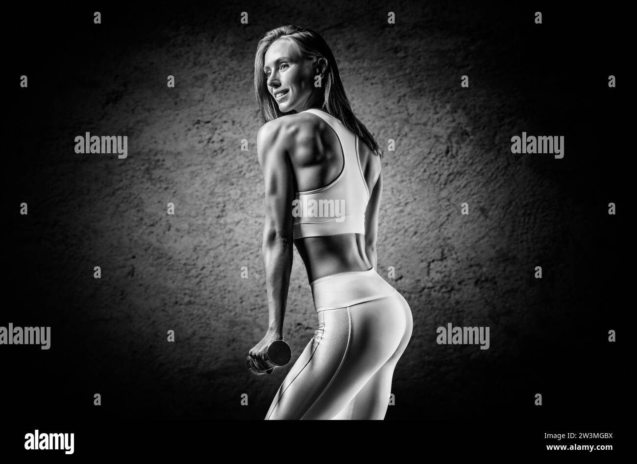 Strong girl is training in the gym with dumbbells. Back view. The concept of bodybuilding, fitness, healthy lifestyle. Light spot. Mixed media Stock Photo