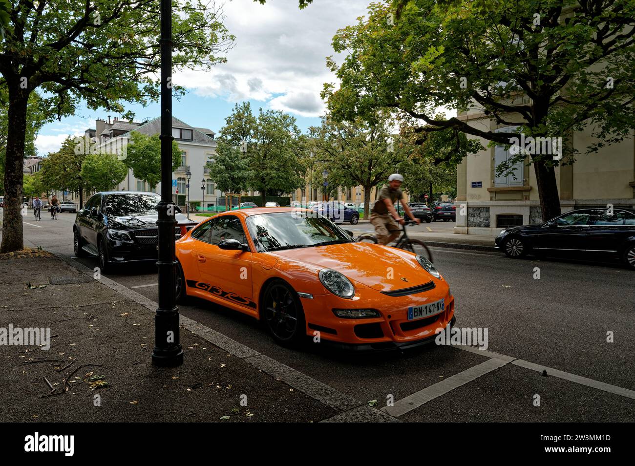 Vibrant orange Porsche 911 GT3 RS parked in a designated spot, surrounded by lush trees and bustling with the energy of swiftly passing cyclists. Stock Photo