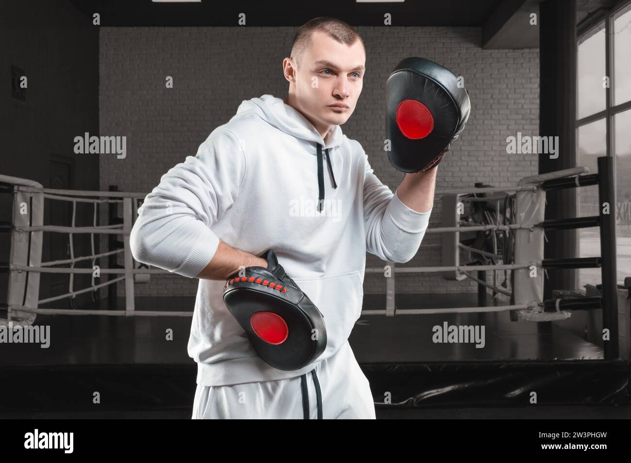 Male trainer in the gym against the background of the ring holds boxing paws. Mixed martial arts