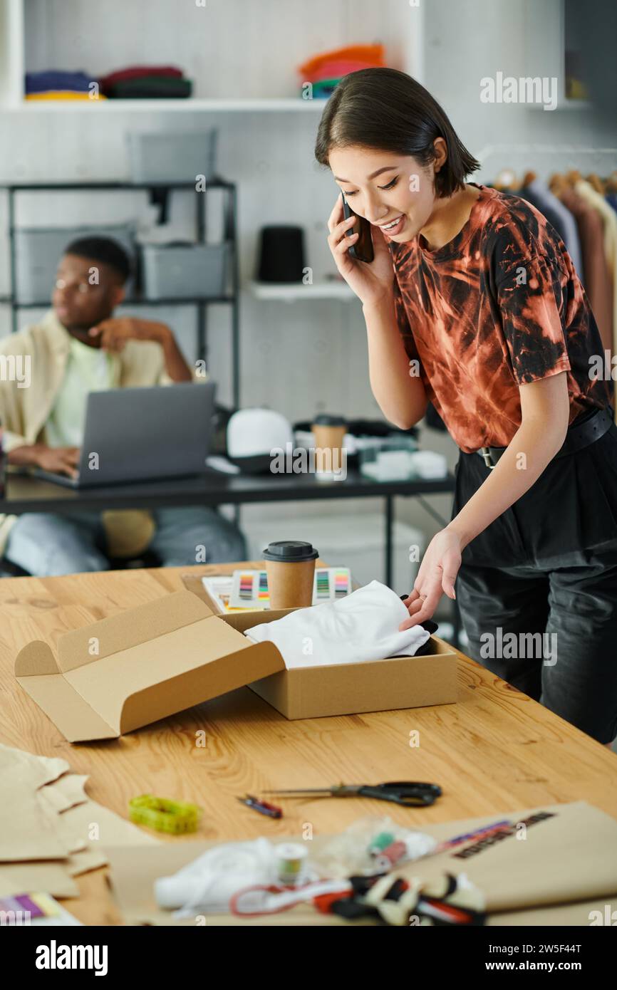 asian designer talking on smartphone and packing garments near african american man in print studio Stock Photo