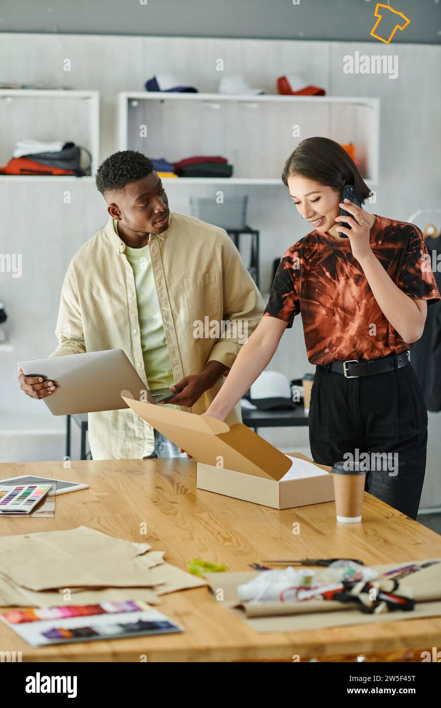 smiling asian woman talking on smartphone near african american man with laptop and carton box Stock Photo