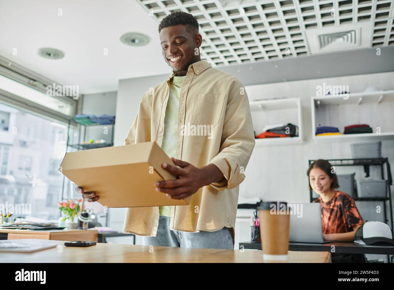 joyful african american man with carton parcel near asian designer at laptop in print studio Stock Photo