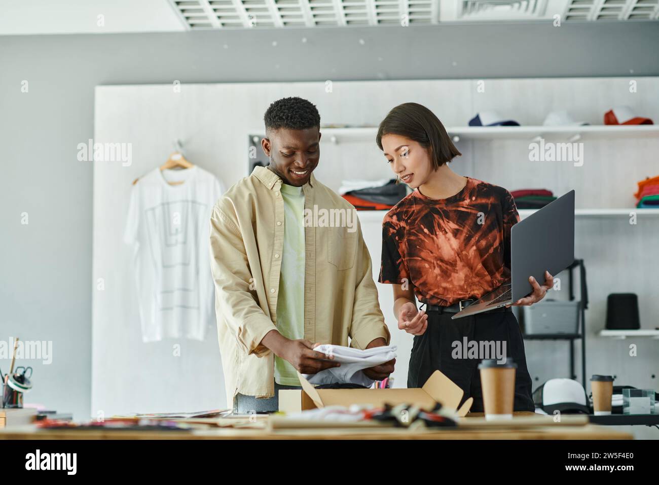 happy african american man packing clothes in box near asian colleague with laptop in print studio Stock Photo