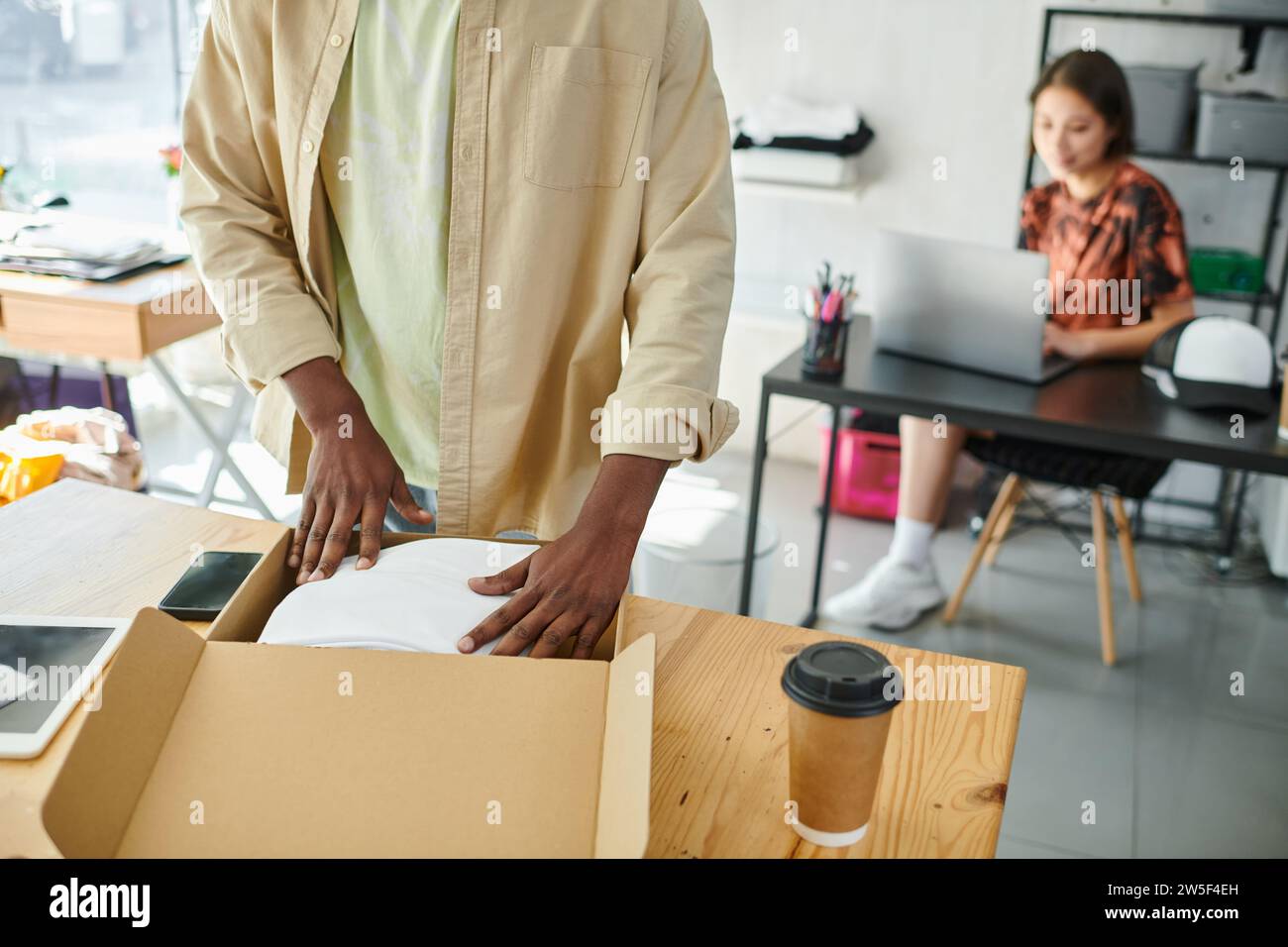 african american man packing clothes in carton box near asian designer at laptop in print studio Stock Photo