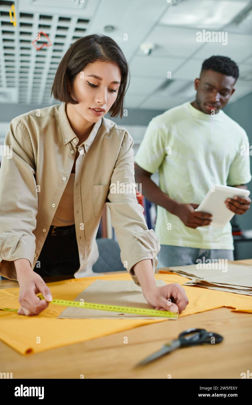 asian fashion designer measuring sewing pattern near african american colleague in print studio Stock Photo