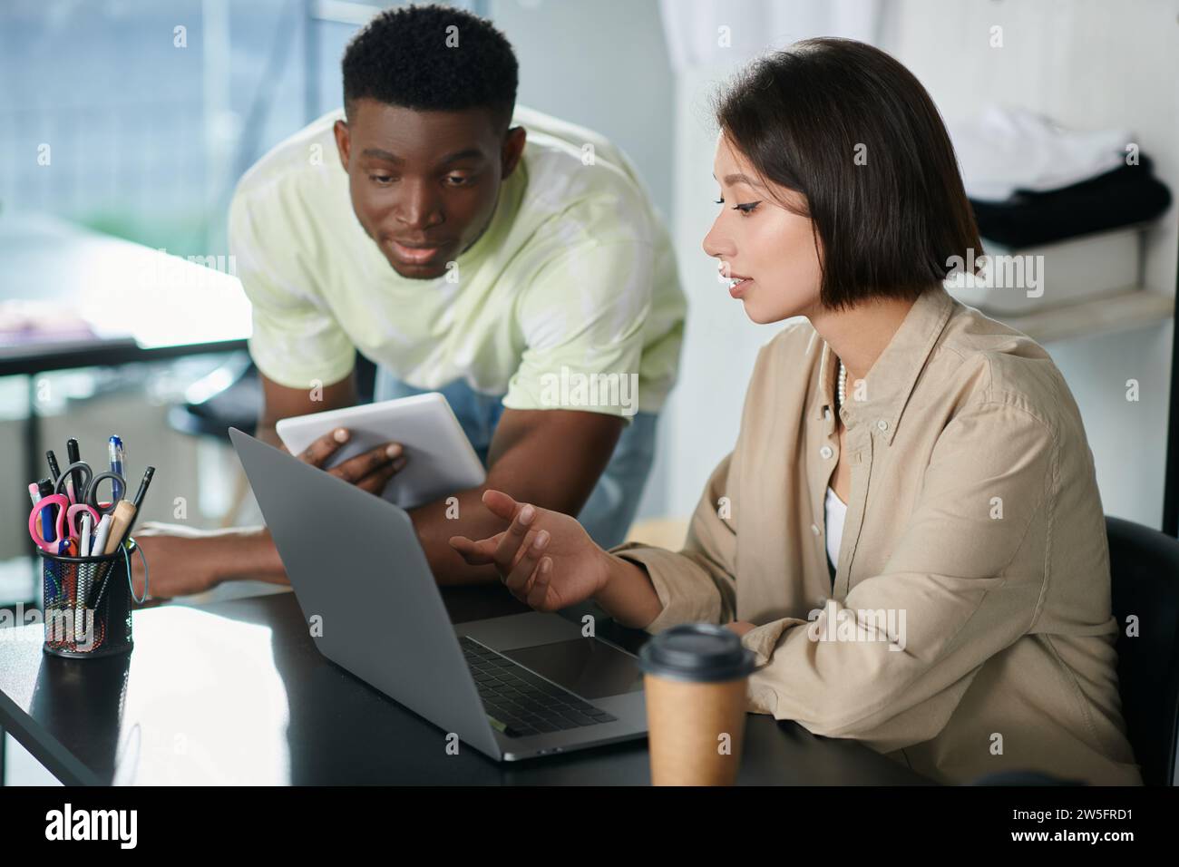 young interracial fashion designer pointing and looking at laptop in print studio, creative teamwork Stock Photo