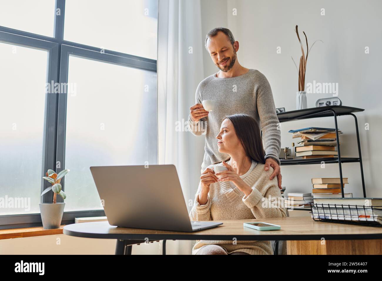 loving husband with coffee cup supporting wife working on laptop at home, unity of child-free couple Stock Photo