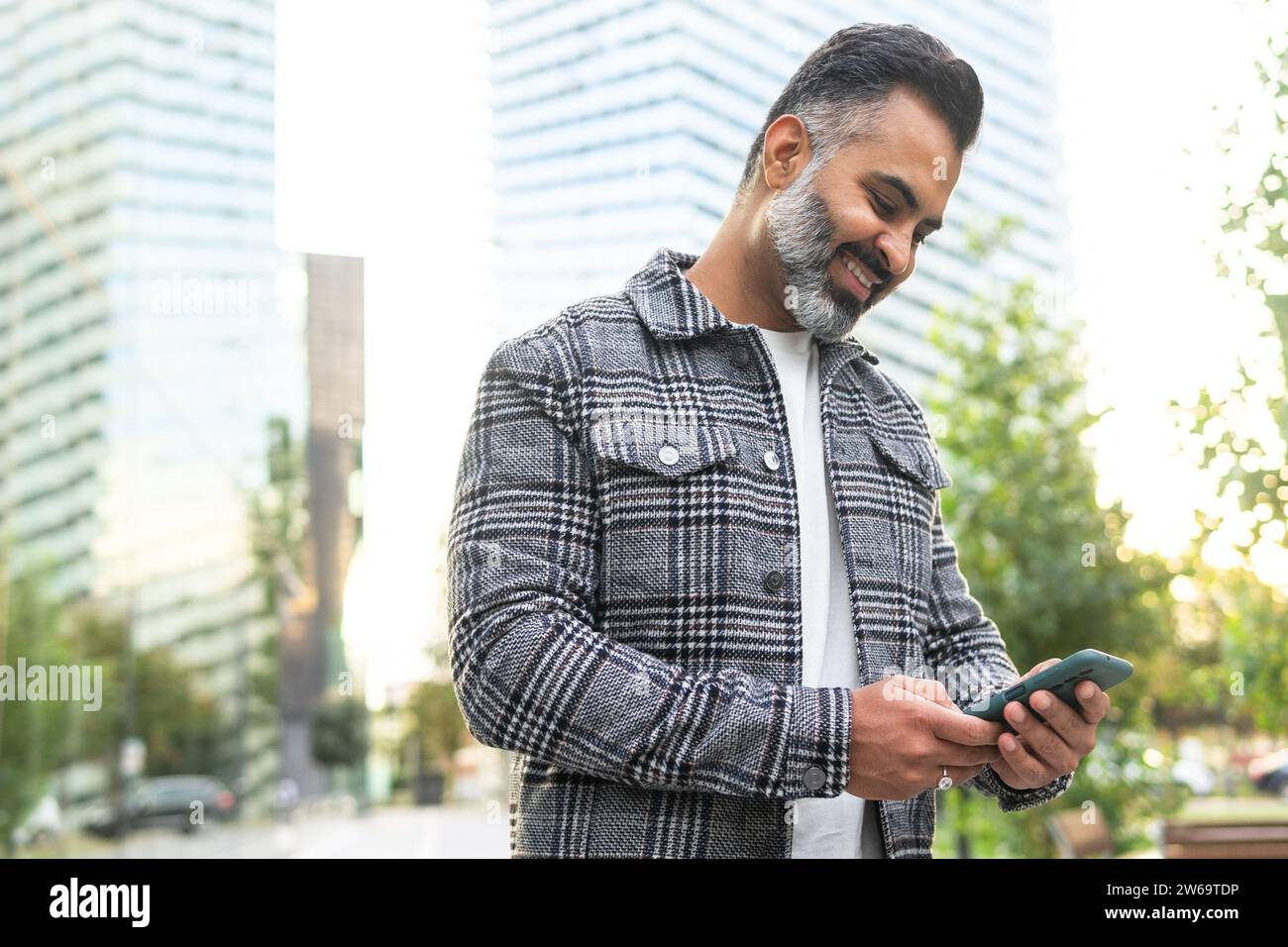 Positive bearded Indian businessman in checkered jacket smiling and browsing smartphone against blurred cityscape background Stock Photo