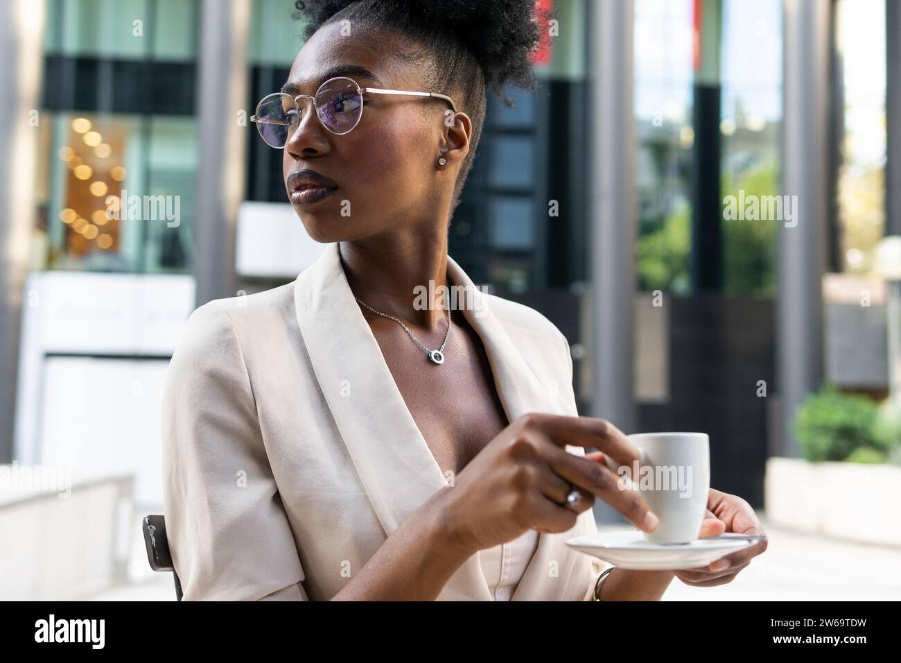 Thoughtful calm African American female professional with eyeglasses and black hair holding coffee cup and looking away while sitting at cafe Stock Photo
