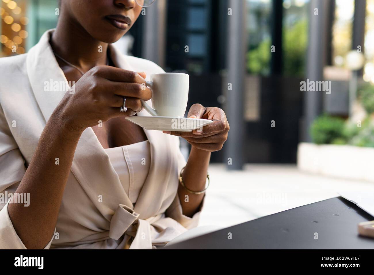 Anonymous crop African American female professional with eyeglasses holding coffee cup while sitting at sidewalk cafe Stock Photo