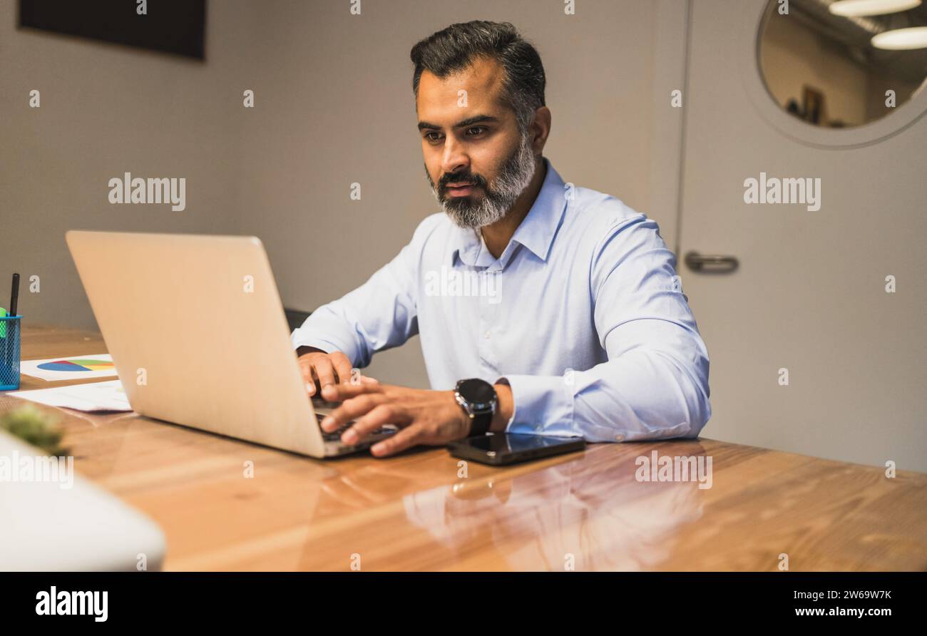 Focused bearded male manager sitting at wooden table and using laptop to work on business project in in modern office Stock Photo