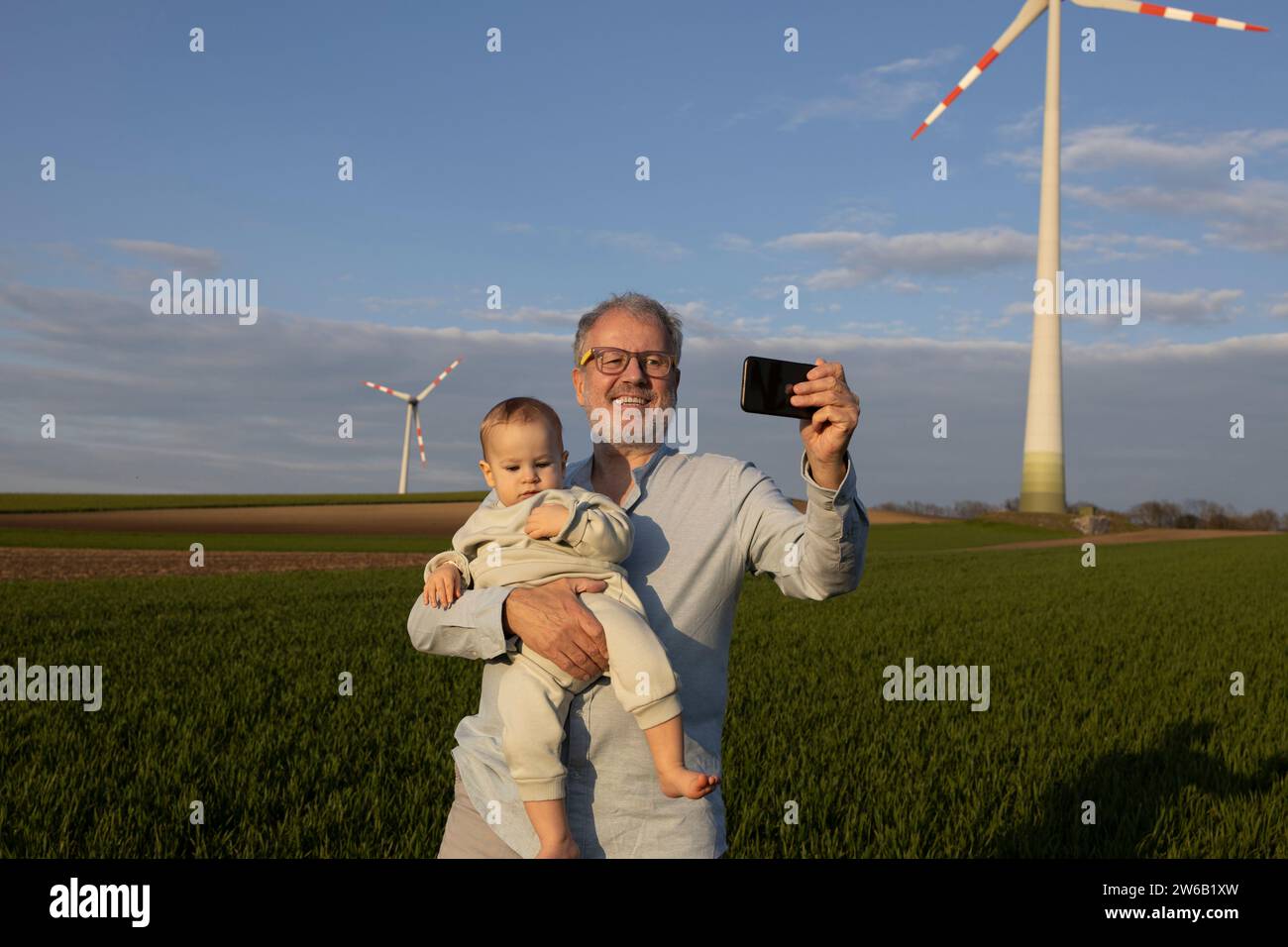 Happy grandfather taking selfie with grandson through smartphone on wind farm against sky Stock Photo