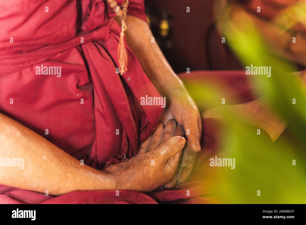 Crop anonymous elderly woman wearing pink Kasaya sitting on floor in lotus pose putting hand on hand while meditating and praying in temple in Thailan Stock Photo