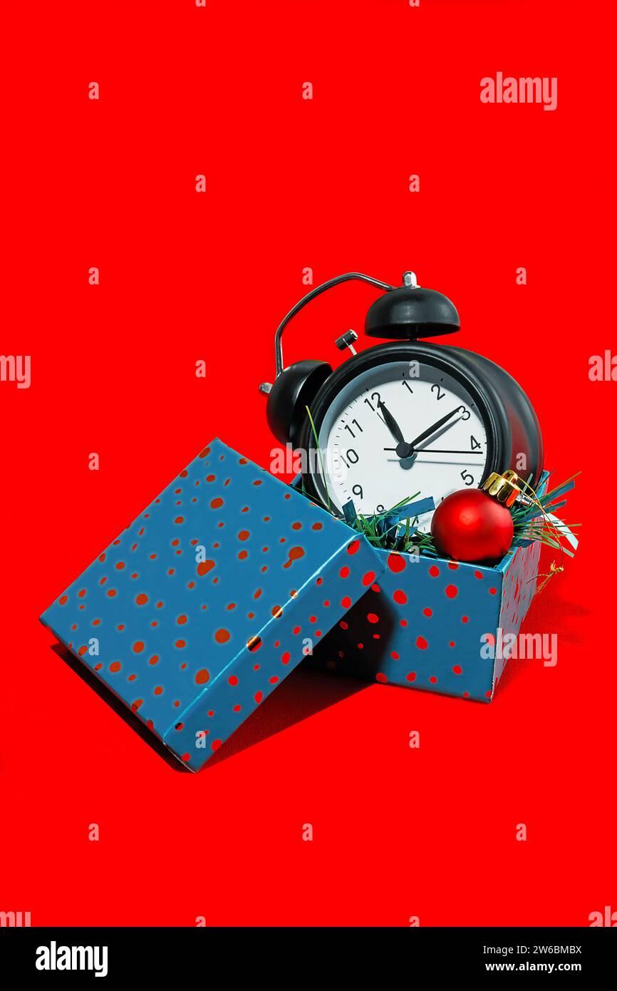 Black alarm clock and red Christmas bauble emerging from a blue gift box set against a vibrant red background. Stock Photo