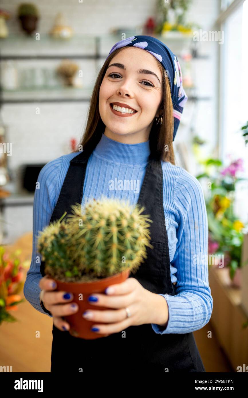 Positive young female gardener wearing headscarf and apron standing in flower shop and holding potted cactus during work Stock Photo