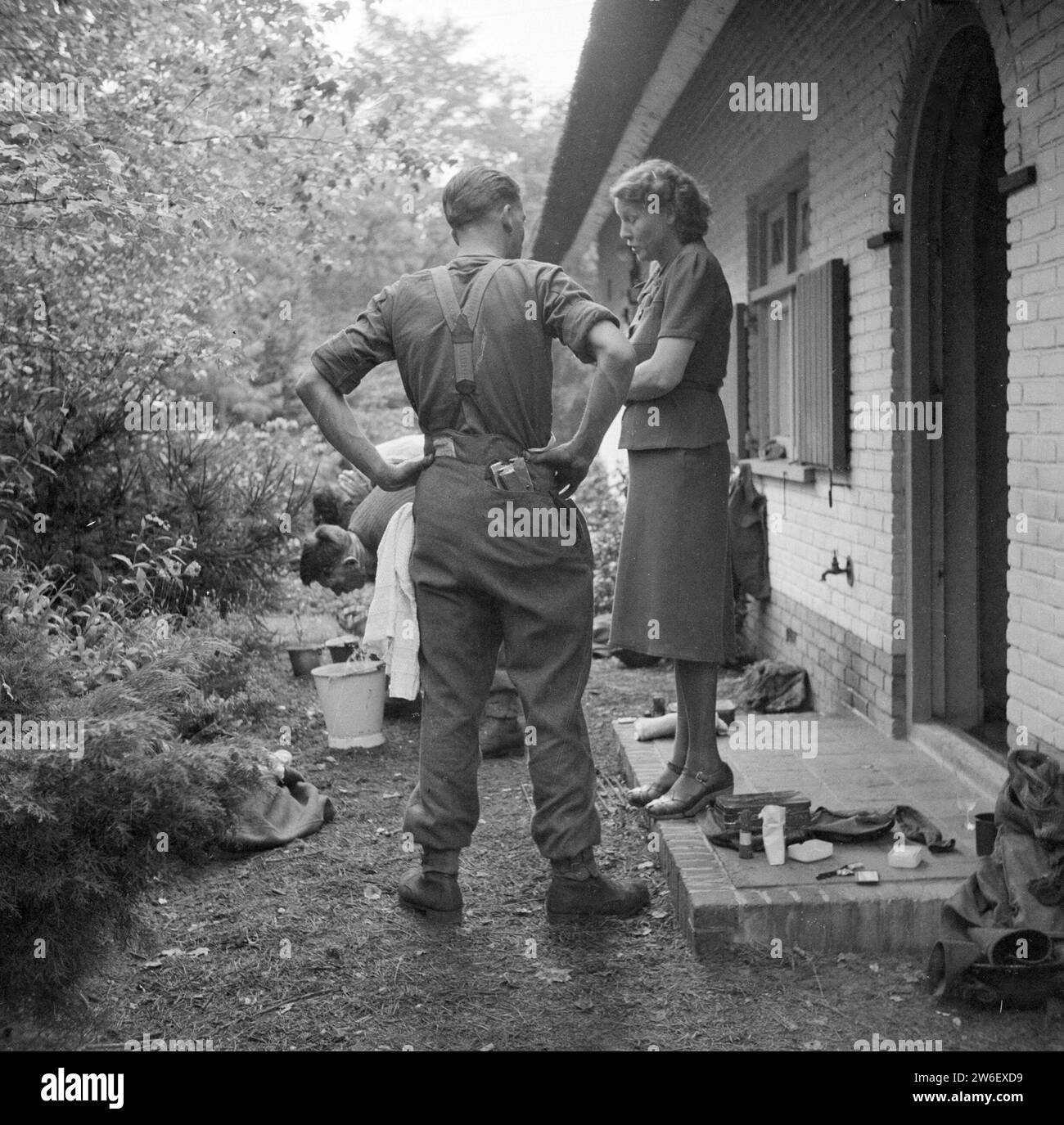 Soldier of the Irish Guards in conversation with a resident of Aalst, with soldiers washing themselves in the background ca. September 18, 1944 Stock Photo