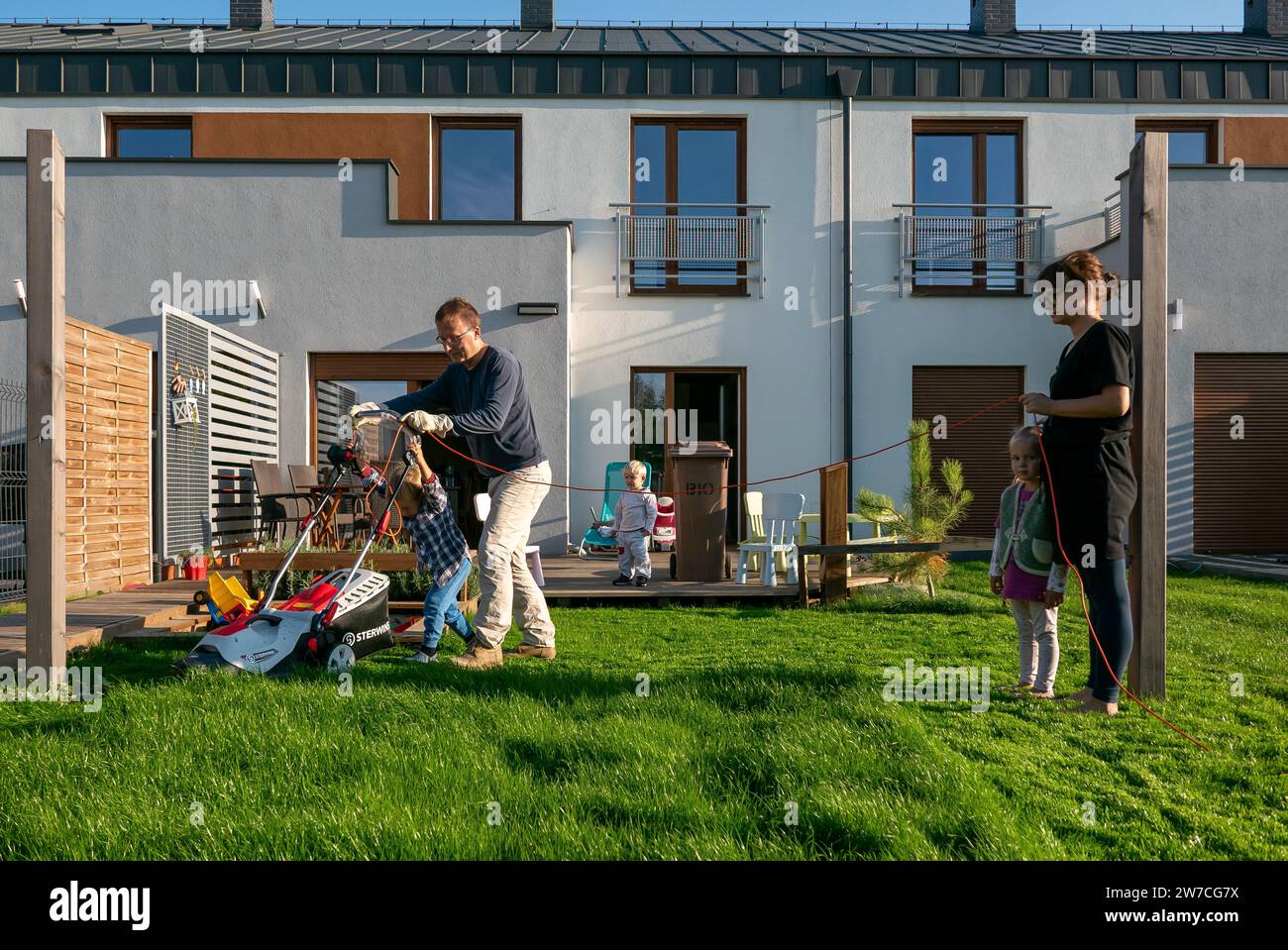 15.09.2018, Poland, Wrzesnia, Wielkopolska - Father and son mowing the lawn in the garden of their terraced house, mother with daughter holding cables Stock Photo