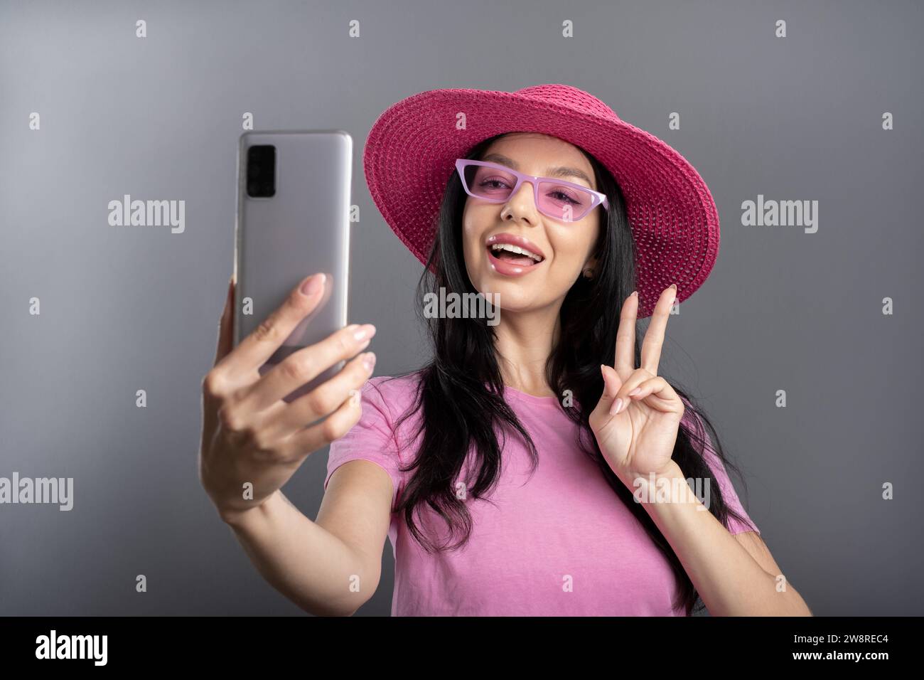 A smiling young woman looking to the camera showing fingers doing victory sign guesture or number two. Wearing a stylish rose-colored summer hat and m Stock Photo