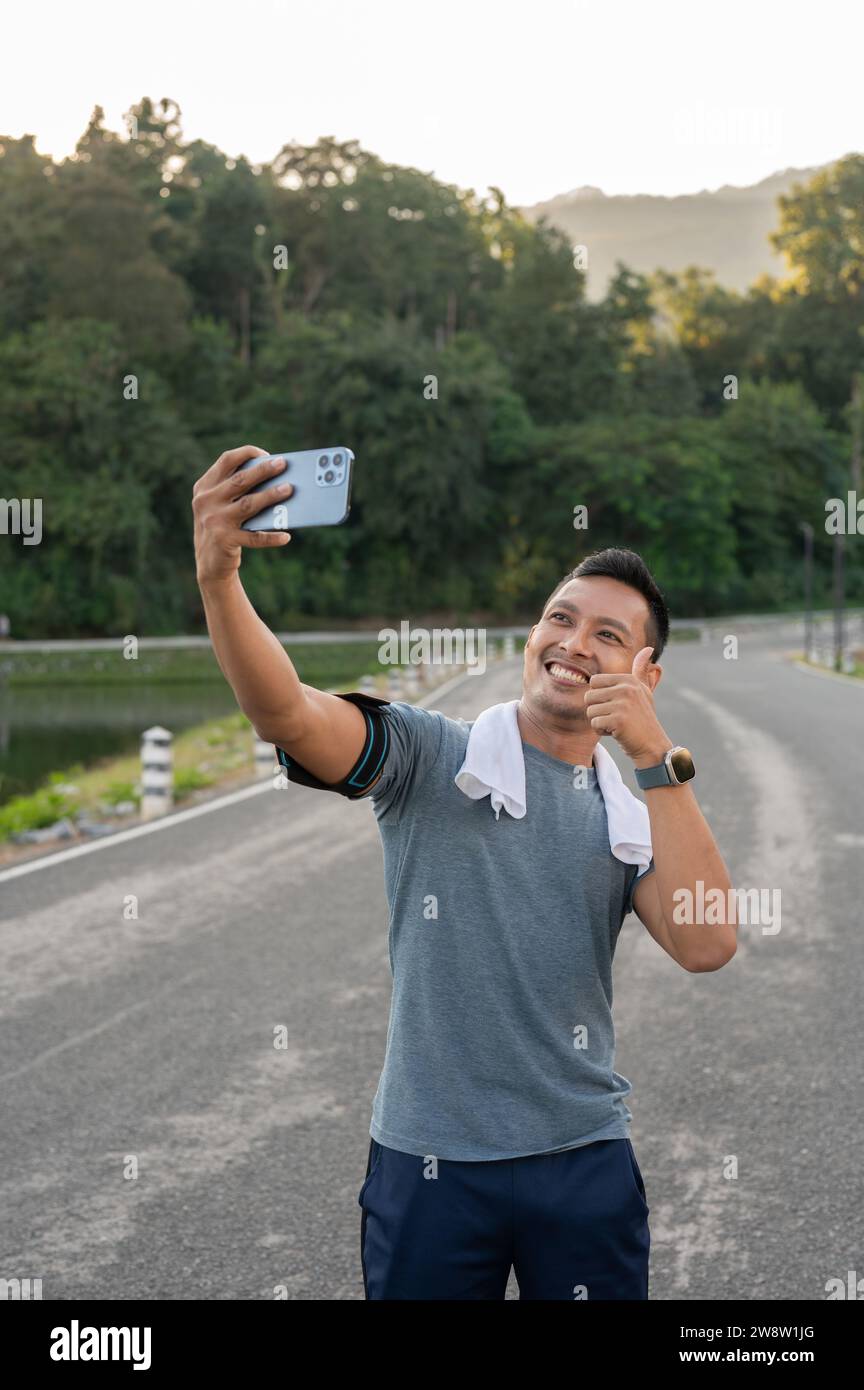 A happy, handsome Asian man in sportswear is showing his thumb up and taking selfie or recording his video during his morning jogging outdoors. Stock Photo