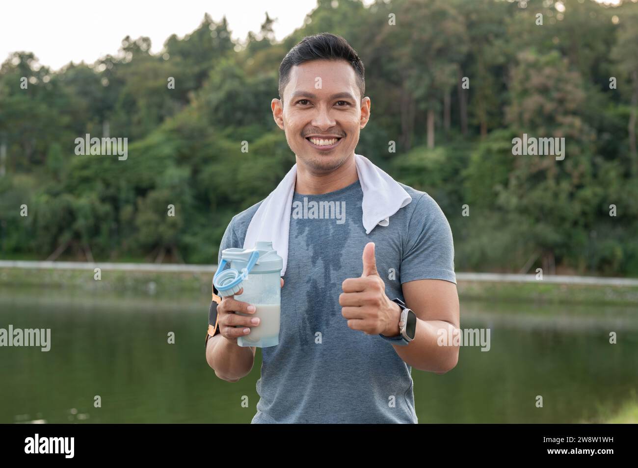 A happy, strong and fit Asian man in sportswear is showing his thumb up and drinking a protein shake after a long run outdoors, having a sport drink s Stock Photo