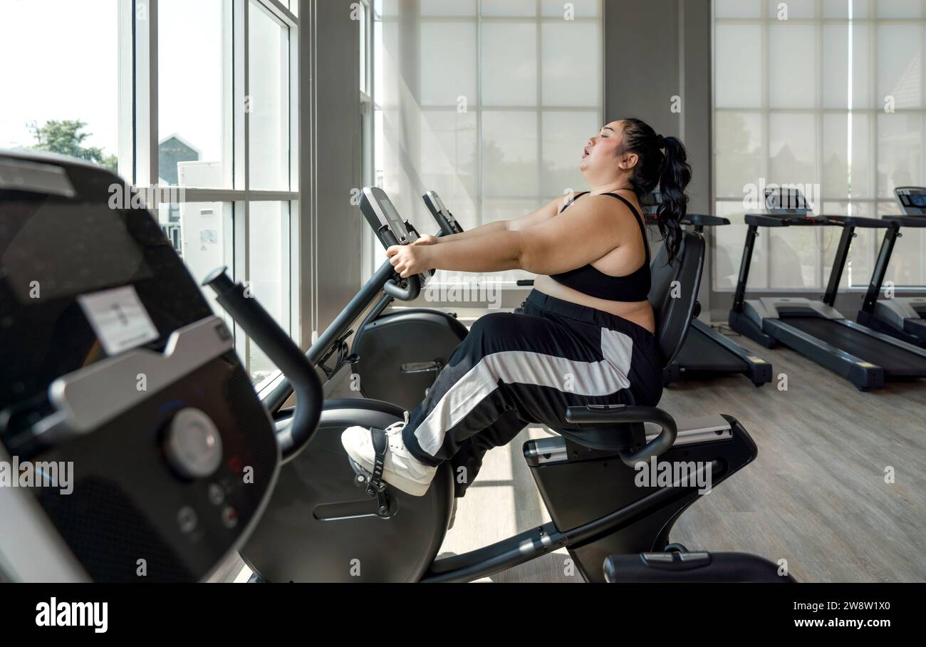 A plus-size lady is energetically riding an exercise bike at a gym, looking focused and determined. Stock Photo