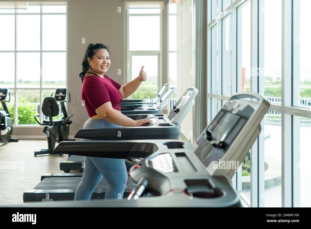 A plus-sized woman is standing on a treadmill in a gym, preparing to start her exercise routine with determination. Stock Photo