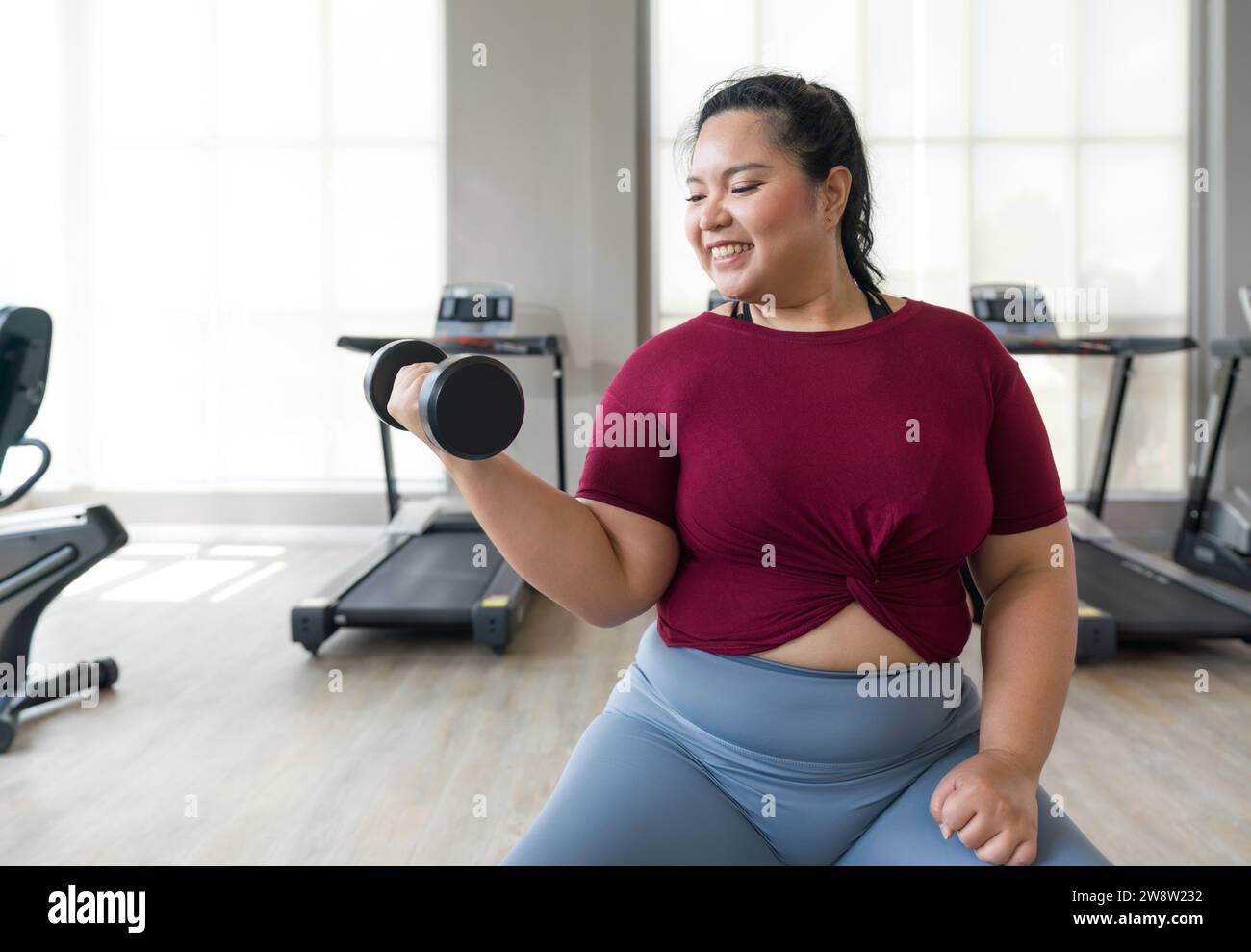 Asian plus-sized woman lifts weights at gym, getting strong and healthy. Stock Photo