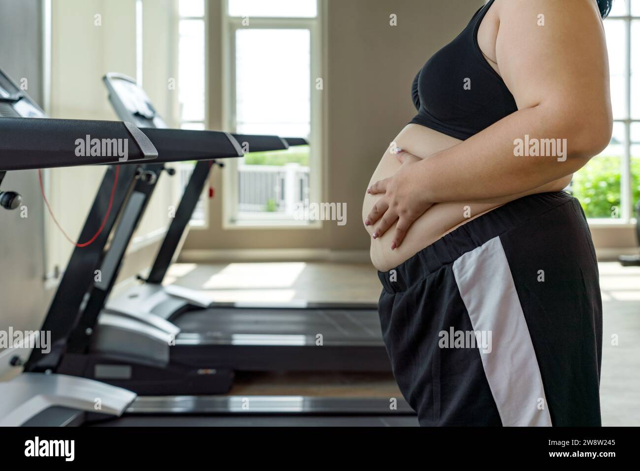 A plus-sized woman placing her hand on her belly, standing confidently in a well-equipped gymnasium. Side view Stock Photo