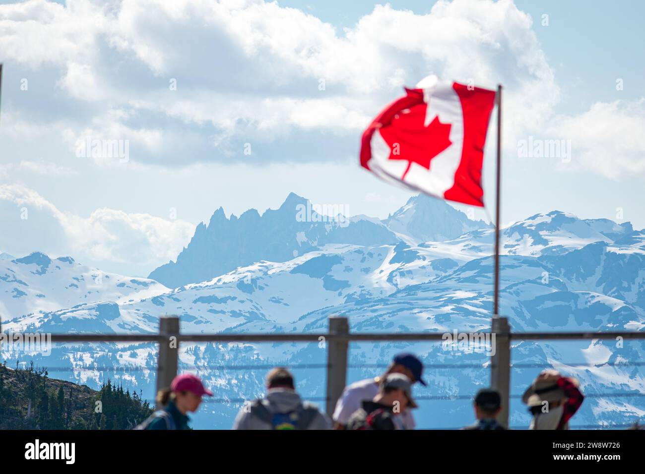 Visitors on Whistler Mountain gaze at the rugged beauty of the Canadian Rockies, with the national flag symbolizing Canada's rich natural heritage. Stock Photo