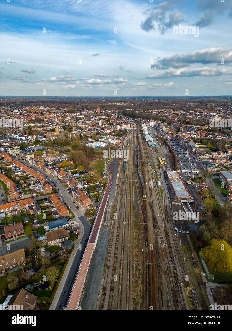 The photograph presents an expansive aerial view over Lier Railway Station in Belgium. The station is a focal point with multiple railway lines converging, surrounded by the urban layout of the city. Residential areas with dense housing, intersected by roadways, are visible, along with patches of greenery providing a contrast to the urban environment. The horizon stretches far, showcasing the town's scale and the open sky above, with scattered clouds casting shadows over the landscape. Aerial View of Lier Railway Station, Belgium. High quality photo Stock Photo