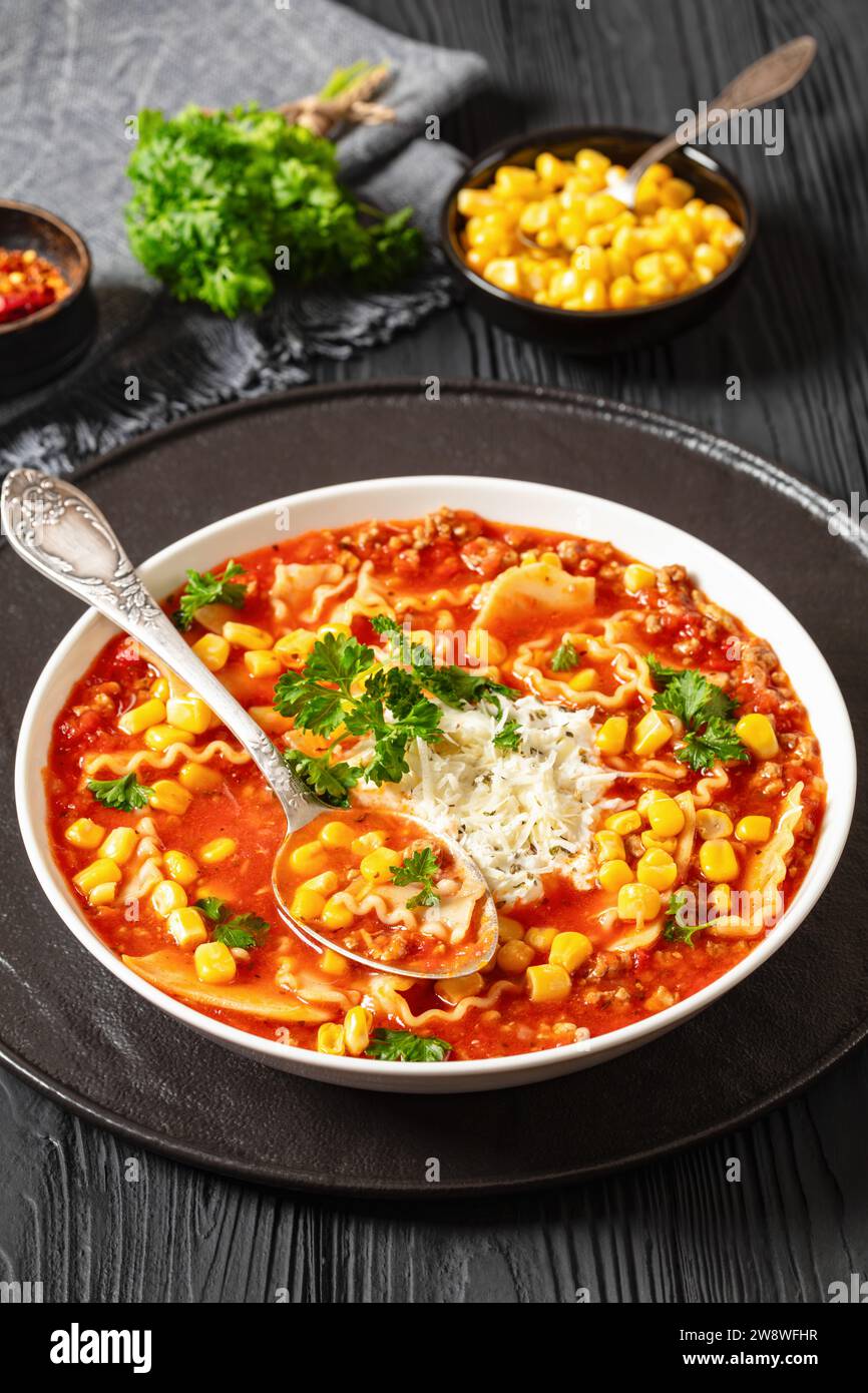 lasagna soup of ground beef, lasagna noodles, tomatoes, corn, spices, ricotta and parmesan cheese in white bowl on black wooden table, vertical view f Stock Photo