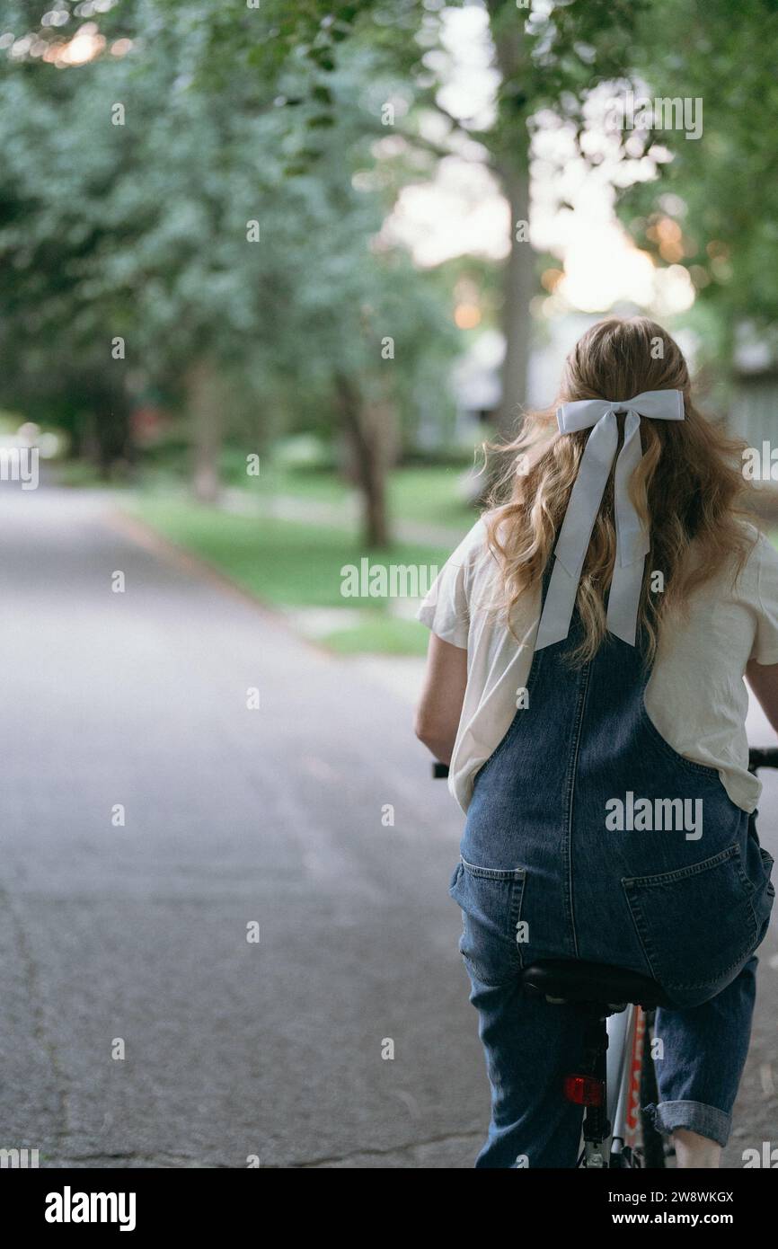 View of the back of a woman riding her bike wearing overalls and a bow Stock Photo