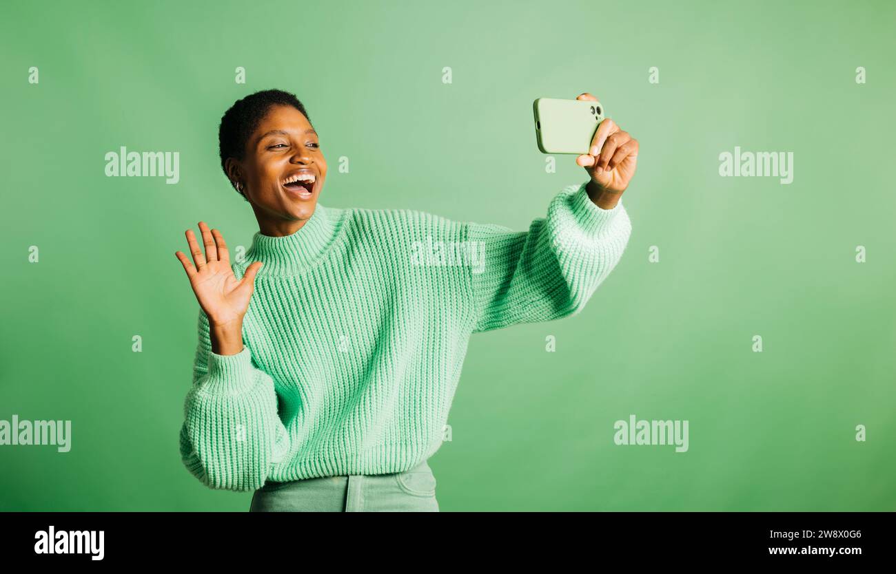 Happy young woman taking a selfie and wearing green clothes in front of a green background in a studio. Woman taking a selfie in a studio. Stock Photo