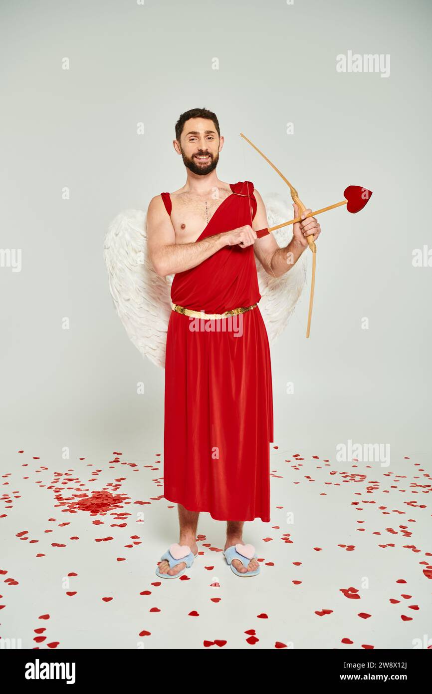 joyful bearded man dressed as cupid archering on grey backdrop, Saint Valentines day costume party Stock Photo