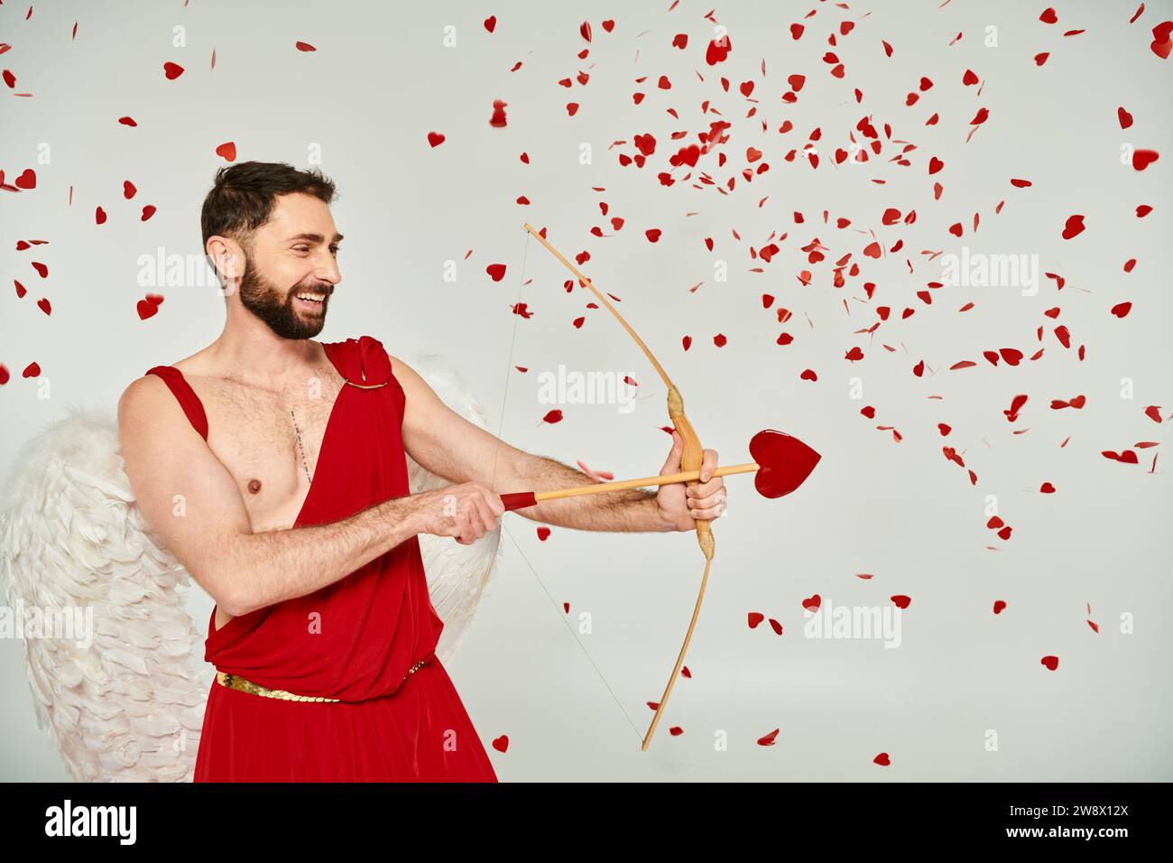 smiling bearded cupid man archering with heart-shaped arrow under red confetti, Saint Valentines day Stock Photo