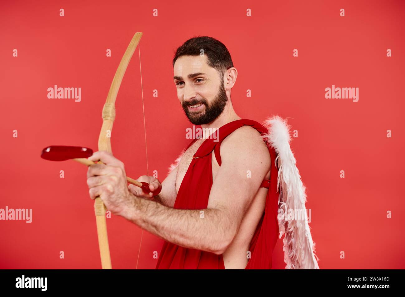 happy bearded man dressed as cupid archering with heart-shaped arrow on red, Saint Valentines day Stock Photo