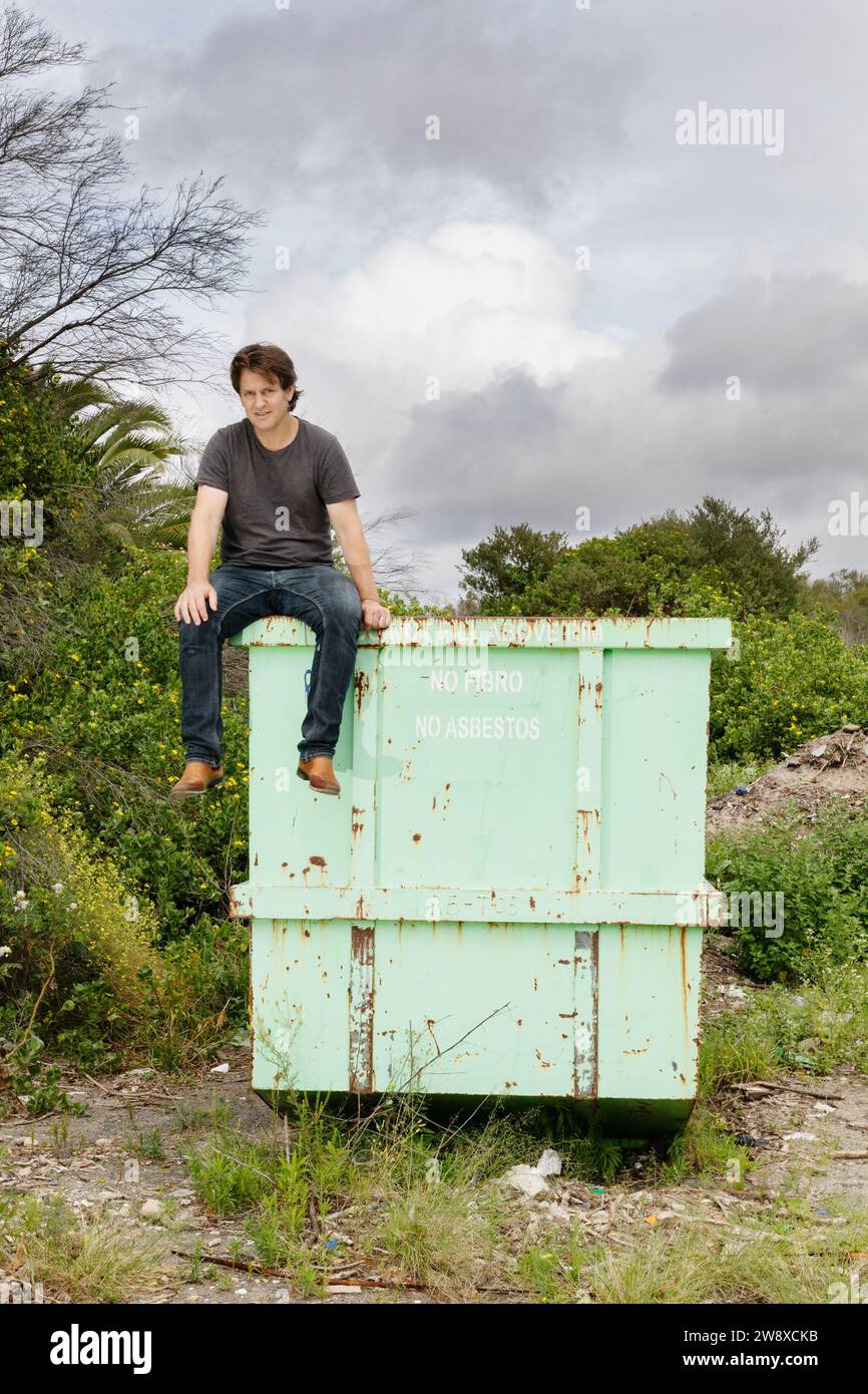 Craig Reucassel, Australian television and radio comedian, known for satirical team The Chaser, and The War on Waste. Sat on metal industrial skip. Stock Photo