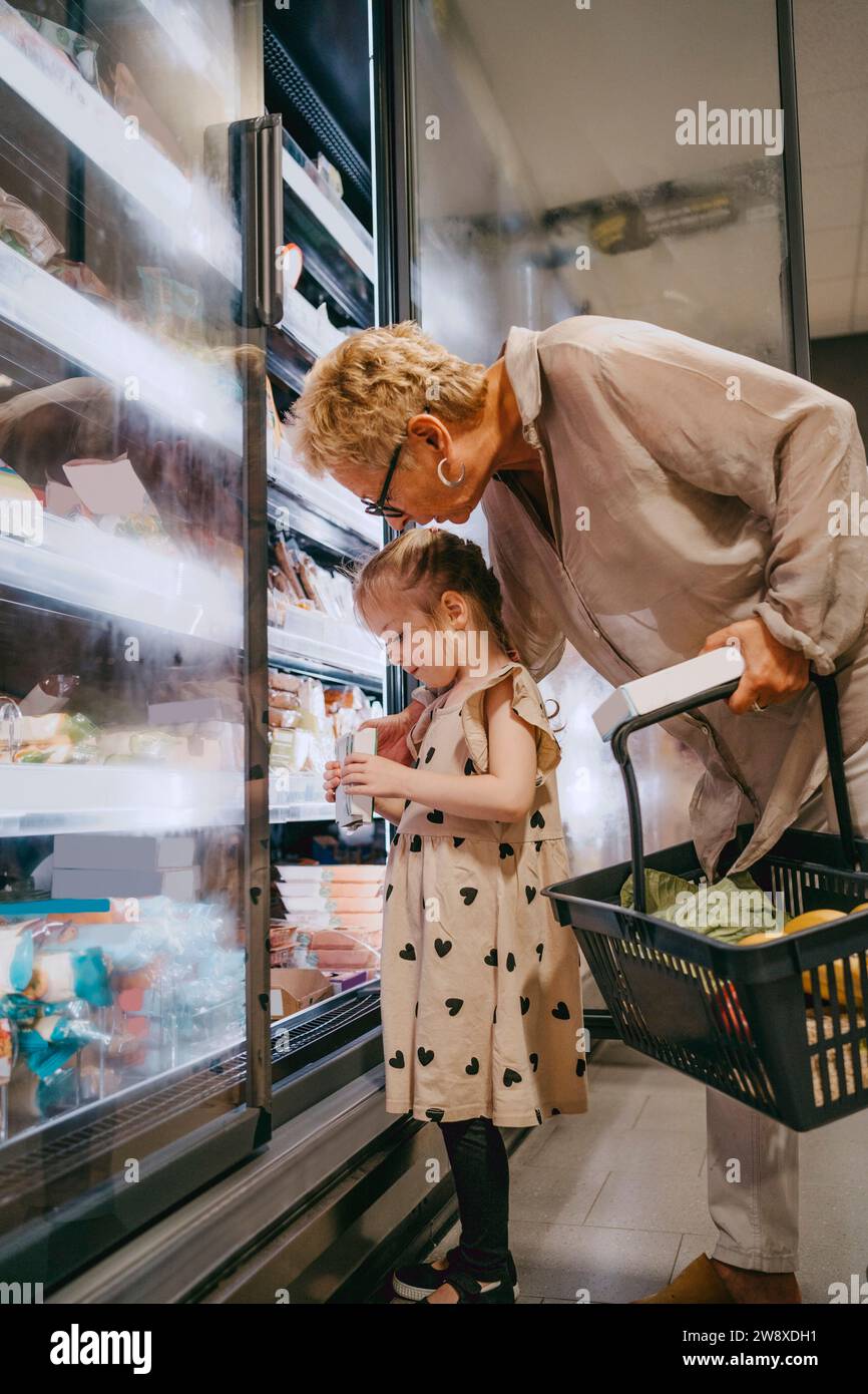 Senior woman with granddaughter choosing frozen food while doing shopping at grocery store Stock Photo
