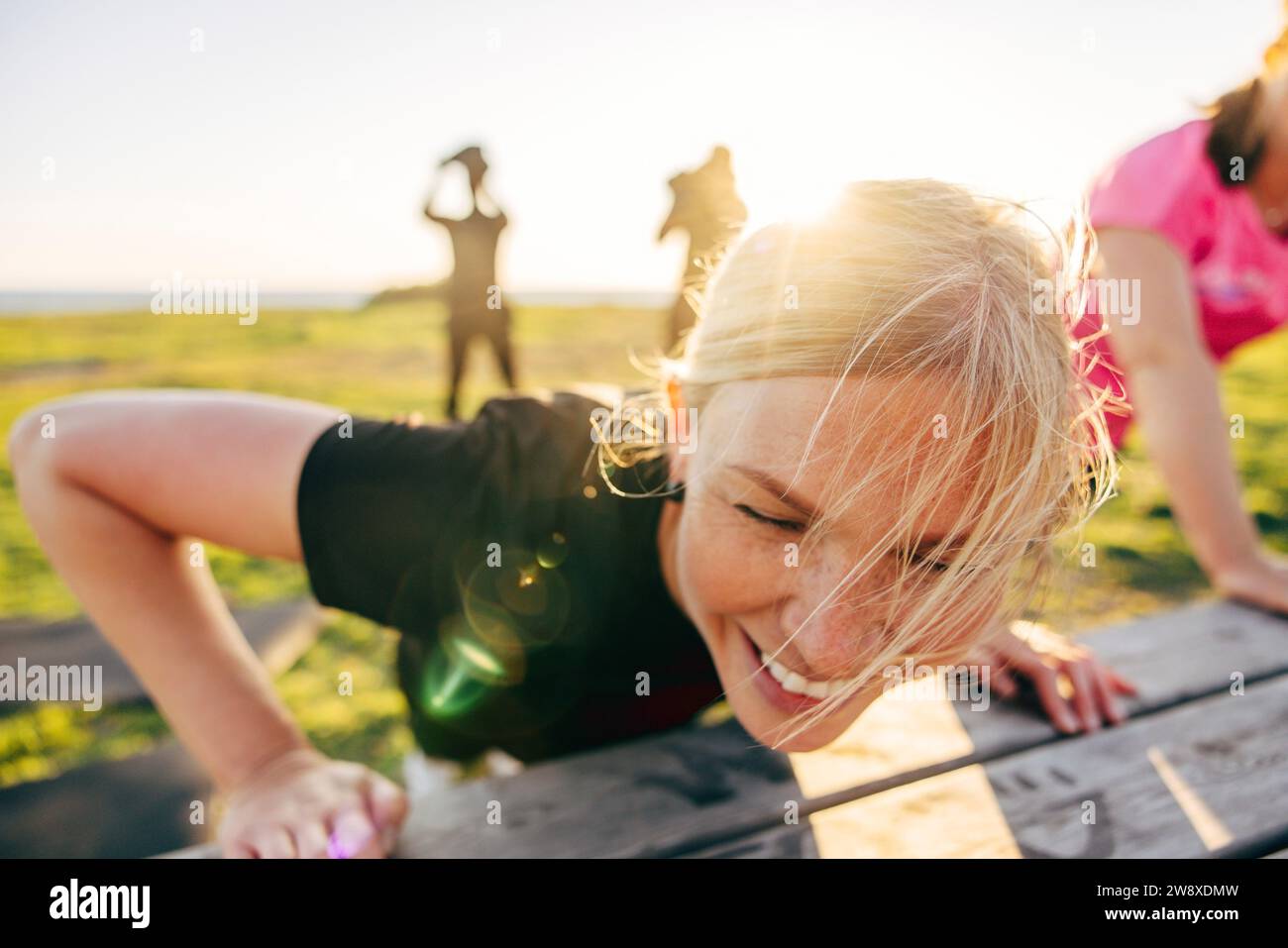 Smiling blond woman doing push-ups on bench during group training at beach Stock Photo