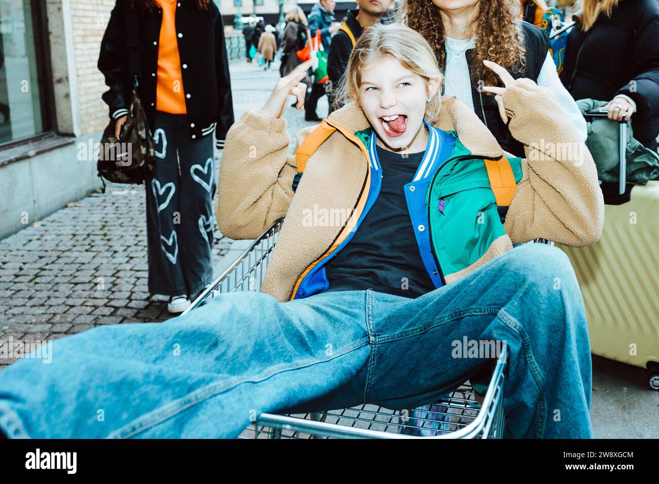 Girl sticking out tongue and gesturing horn sign sitting on shopping cart at footpath Stock Photo