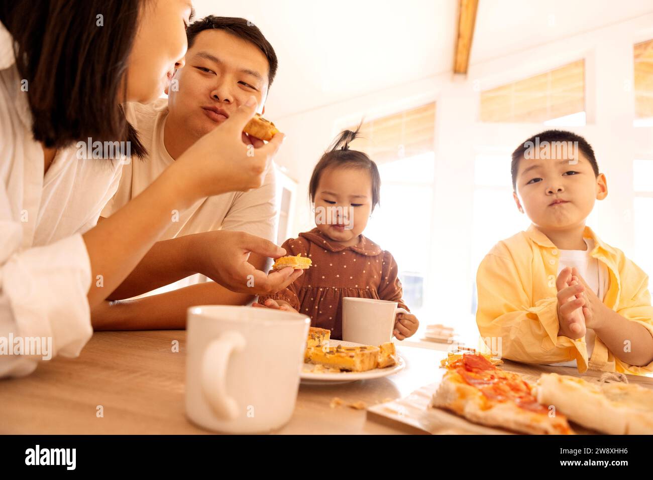A happy Asian family is eating pizza in the kitchen at home. A young Korean couple and their cute children are having lunch. Dad, mom, daughter and so Stock Photo