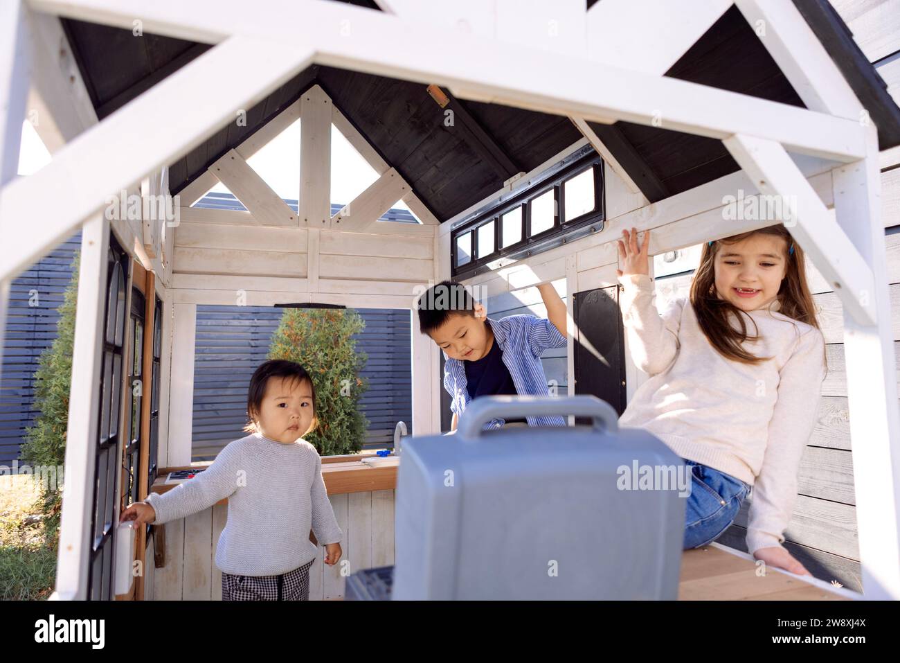 Three small children of different races are playing in a wooden children house. Asian and Caucasian ethnic cute kids have fun together in the backyard Stock Photo