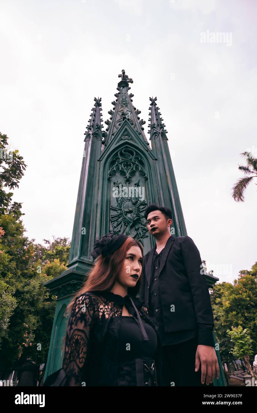 a couple of Asian teens standing near a spooky cemetery full of green trees in the afternoon Stock Photo